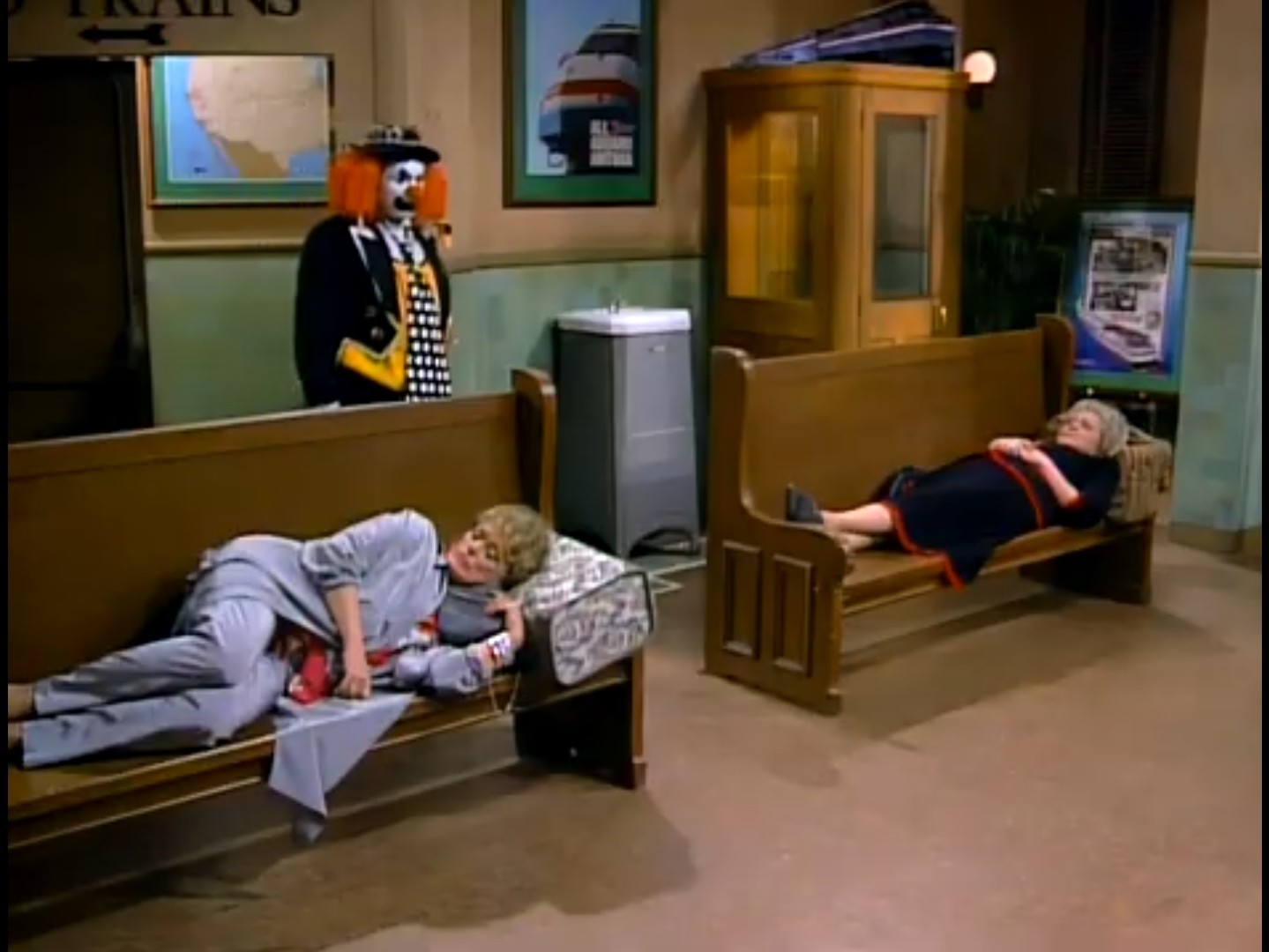 Blanche and Rose sleeping in a train station as a clown walks in